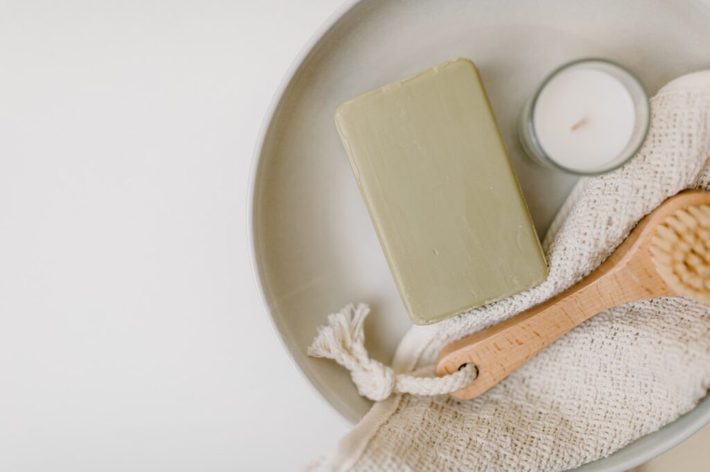A tray of spa tools including an wooden exfoliating brush, an exfoliation mit, a green bar of soap and a green tray to hold it all.
